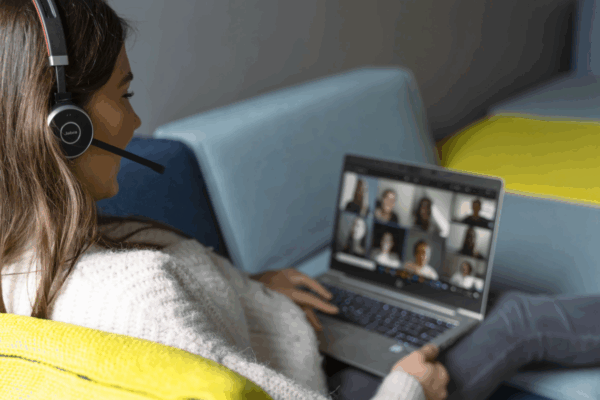 Junge Mitarbeiterin im Online-Meeting mit Headset, Laptop auf dem Schoß – interaktives Lernen, Teamkommunikation und modernes hybrides Arbeiten.