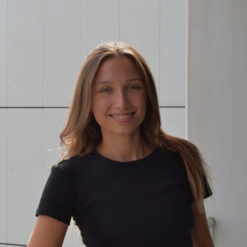 Mitarbeiterin mit brünetten Haaren lächelt auf Terrasse – wirkt spontan und sympathisch, symbolisiert zugängliche Unternehmenskultur.